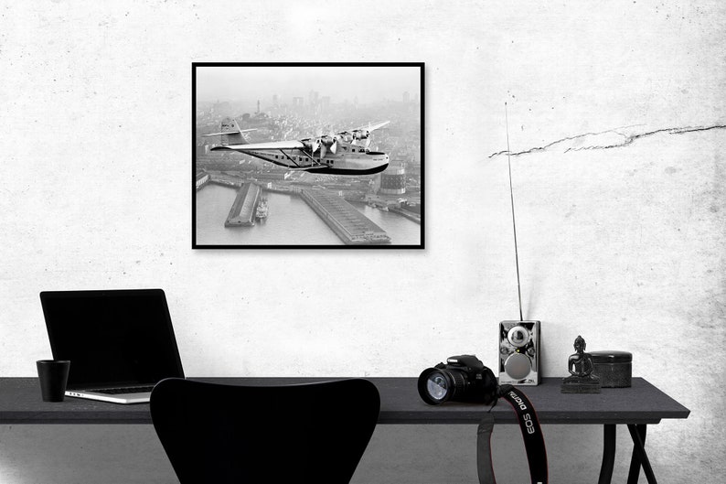 May include: Black and white photograph of a vintage seaplane flying over a city. The plane is in mid-air, with the city skyline visible in the background. The photo is framed in a black frame.