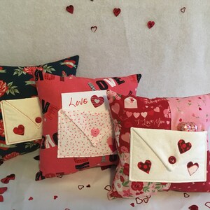 May include: Three decorative pillows with a Valentine's Day theme. Each pillow has a white envelope pocket with a red heart on the front. The pillows are made of different fabrics with various patterns, including roses, hearts, and polka dots. The pillows are arranged on a white surface with red hearts scattered around them.