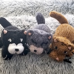 May include: Three handmade knitted cat toys in black, gray, and brown. Each cat has a unique design with pink accents on the ears and a white muzzle. The toys are lying on a soft, gray, fluffy surface.
