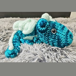 May include: A teal blue and light green crocheted dragon toy with a single black eye. The dragon is lying on a gray fluffy surface.