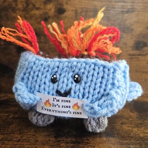 May include: A blue knitted campfire with a smiling face and orange yarn flames. The campfire is holding a sign that reads "I'm fine, It's fine, Everything's fine."