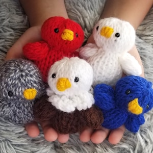 Peut inclure: Cinq jouets d'oiseaux en tricot de différentes couleurs. Les oiseaux sont rouge, blanc, gris, marron et bleu. Ils ont des yeux noirs et des becs jaunes.
