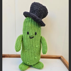 May include: A knitted green cactus toy wearing a dark grey top hat. The cactus has a smiling face with black eyes and a curved mouth. The toy has arms and feet, and is made of a textured yarn.