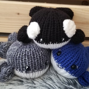 May include: Three handmade, knitted orca whale plushies. One is black and white, one is blue and white, and the third is gray. Each has black button eyes and a smiling face. The plushies are on a gray, fuzzy surface with a wooden background.