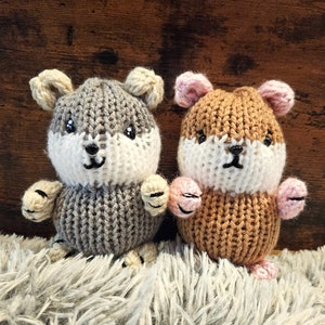 May include: Two knitted hamster toys, one gray and one brown, with white faces and pink ears. The toys are sitting on a fluffy white surface.