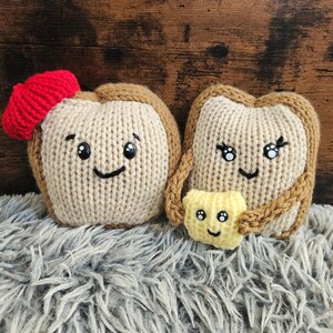 May include: Hand-knitted toast-shaped plush toys with smiling faces. One slice has a red bow, the other has eyelashes. A small, smiling butter square is held between them. The toys are on a gray, fluffy surface.