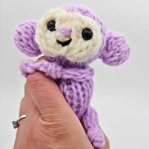 May include: A knitted monkey doll in lavender and cream. The monkey has a smiling face with black eyes and a cream-coloured face. The doll is held in a hand, showing the intricate knit details and soft texture of the toy. A charming handmade item.