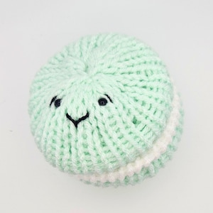Könnte beinhalten: Ein mintgrünes, gestricktes Spielzeug in Form eines Macarons mit einem lächelnden Gesicht. Das Spielzeug hat eine weiße Füllung und schwarz gestickte Augen und Mund. Die Strickstruktur ist im gesamten Spielzeug sichtbar.