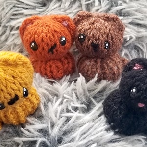 Puede incluir: Cuatro pequeños juguetes de peluche tejidos en forma de osos. Los osos son naranja, marrón, amarillo y negro. Tienen ojos y narices bordados.