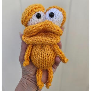 May include: Handmade orange knitted duck toy with large white and black eyes, a matching hat, and a scarf. The duck has small legs and arms. The toy is made of soft yarn and is a unique, handcrafted item.