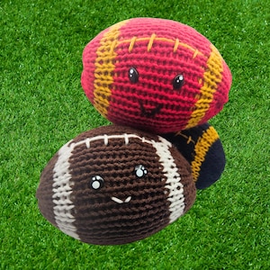 May include: Two crocheted footballs with smiling faces rest on green grass. The top ball is red with yellow stitching, while the bottom one is brown with white stripes. Both have black eyes and stitched smiles, creating a playful, handmade look.