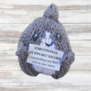 May include: A gray knitted shark plushie with a white belly and a small fin. The shark holds a sign that reads "EMOTIONAL SUPPORT SHARK - reminding you that you're jaw-some."