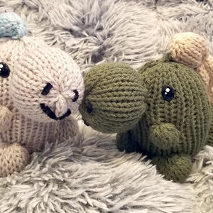 May include: Two knitted dinosaur plush toys. One is light beige with a blue crest, the other is olive green with a tan crest. Both have black button eyes and stitched smiles. They are sitting on a fluffy, gray surface.