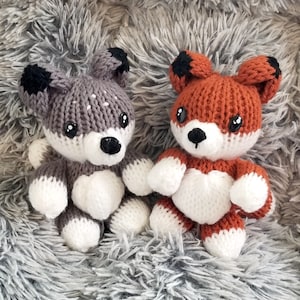 May include: Two knitted stuffed animals, a gray wolf and an orange fox, are sitting on a gray fluffy surface. Both animals have white faces and black eyes. They are holding white hearts in their paws.