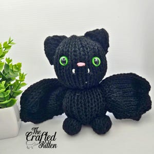 May include: A handmade black knitted bat with green eyes, a pink nose, and small white fangs. The bat has large, spread wings and small feet. The Crafted Kitten logo is visible in the lower left corner.