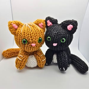 Puede incluir: Dos juguetes de gato tejidos. Uno es naranja con detalles blancos y beige, orejas internas rosas y ojos verdes. El otro es negro con orejas internas rosas, detalles blancos y ojos verdes. Ambos tienen vientre blanco.