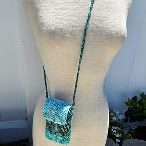 May include: A small, teal and green knitted crossbody bag with a long strap. The bag is made of a soft, textured yarn and has a flap closure.