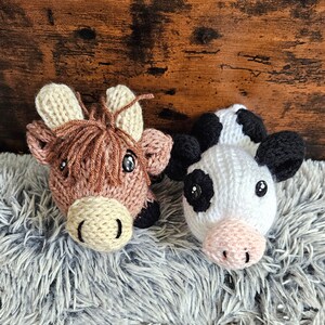 Puede incluir: Dos cabezas de animales tejidas a mano: una vaca de las Tierras Altas y una vaca. La vaca de las Tierras Altas es marrón con detalles beige, la vaca es blanca con manchas negras. Ambas tienen ojos grandes y negros.