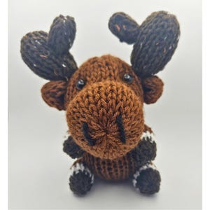 May include: A handmade brown knitted moose toy with dark brown antlers and black eyes. The moose has a textured appearance and is sitting upright. The hooves have white and black accents. The toy is made of yarn.