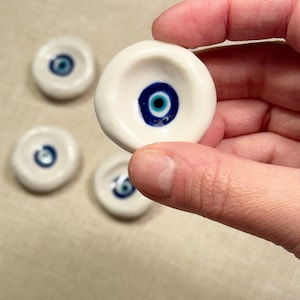 May include: White ceramic eye-shaped beads with a blue and black iris design. The beads are round with a slight indentation in the center. One bead is held in a hand, with three others visible in the background.