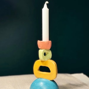 Puede incluir: Un portavelas de cerámica apilado y colorido con una vela blanca. La base es azul, seguida de un diseño geométrico amarillo, verde y naranja. Un artículo de decoración del hogar único.