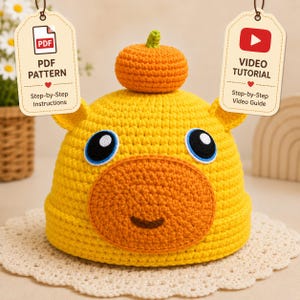 May include: A handmade yellow crochet hat with an orange face and a small pumpkin on top. The hat has blue and black eyes and small ears. The image includes tags that say "PDF Pattern" and "Video Tutorial".