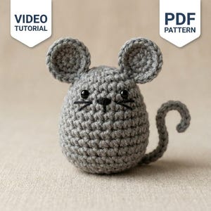 Puede incluir: Un ratón de ganchillo gris con orejas redondas, ojos de cuentas negras y bigotes bordados. El ratón tiene una cola curva y un cuerpo redondeado. Dos pancartas blancas en la parte superior muestran el texto VIDEO TUTORIAL y PDF PATTERN.