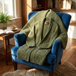 May include: An olive green knit cardigan with a shawl collar and a textured cable knit pattern. The garment features long sleeves, two front pockets, and a small rectangular brand label sewn into the inner neckline area of the collar.