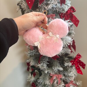 May include: Pink fuzzy keychain with a gold ring and two white bows. The keychain is shaped like a mouse head.