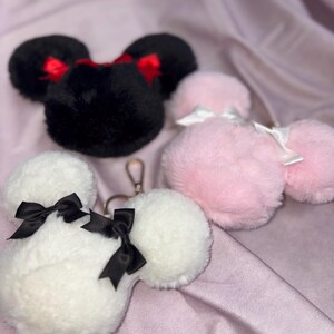 May include: Three plush keychains shaped like Mickey Mouse ears. One is black with red bows, one is pink with white bows, and one is white with black bows.