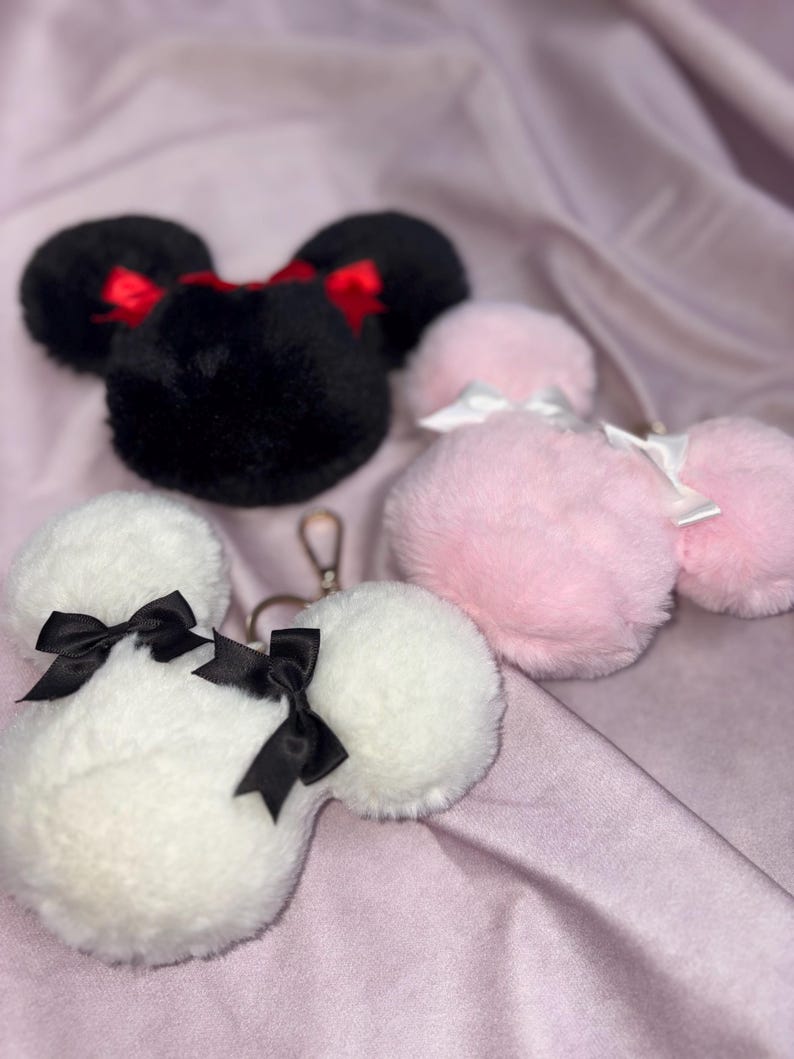 May include: Three plush keychains shaped like Mickey Mouse ears. One is black with red bows, one is pink with white bows, and one is white with black bows.