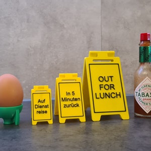 Puede incluir: Un huevo marrón en un huevera verde, tres letreros amarillos con texto negro y una botella de salsa Tabasco. Los letreros dicen "Auf Dienstreise", "In 5 Minuten zurück" y "OUT FOR LUNCH".