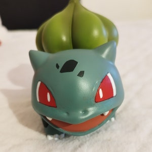 Può includere: Figurina Pokemon verde e blu di Bulbasaur con occhi rossi. La figurina è in plastica e ha un design dettagliato.