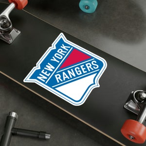 Op de afbeelding: Een zwart skateboard met een witte sticker van het New York Rangers logo. Het logo is blauw, rood en wit met de woorden "New York Rangers" in een witte omtrek.