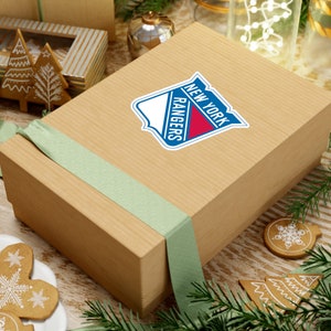 Op de afbeelding: Een bruine cadeaudoos met een groen lint en een sticker met het logo van de New York Rangers. De doos staat op een tafel met een kerstthema.