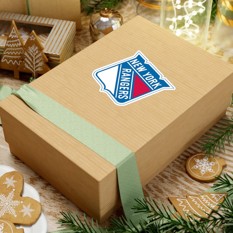 Op de afbeelding: Een bruine cadeaudoos met een groen lint en een sticker met het logo van de New York Rangers. De doos staat op een tafel met een kerstthema.
