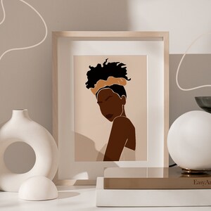 May include: A framed illustration of a woman with dark skin, wearing a brown headband and a white top. The background is a light brown colour. The frame is light wood.