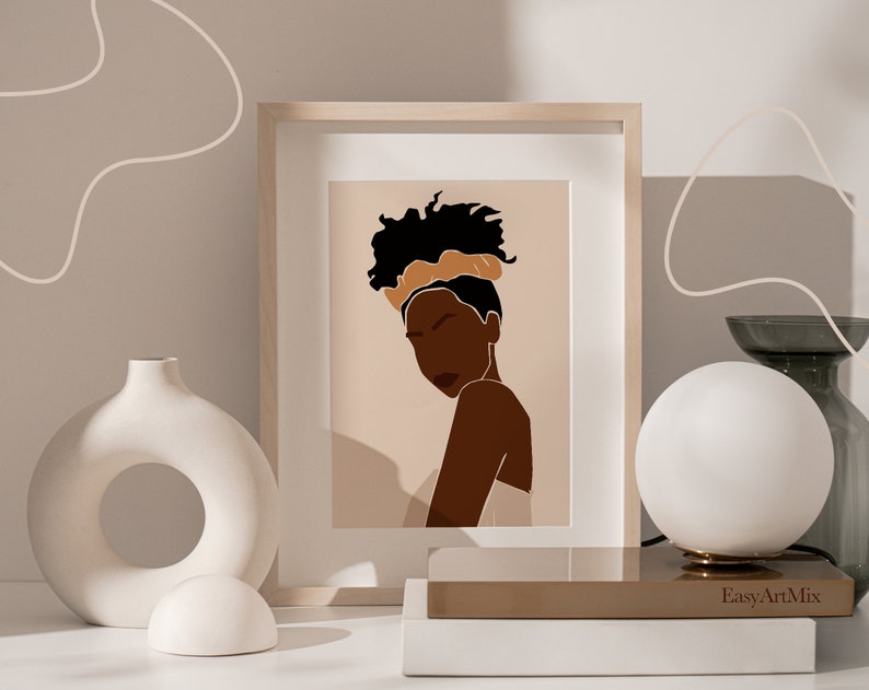May include: A framed illustration of a woman with dark skin, wearing a brown headband and a white top. The background is a light brown colour. The frame is light wood.