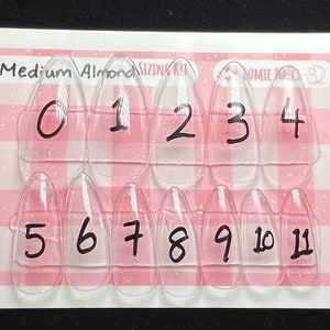 May include: A set of 11 clear plastic nail tips in a medium almond shape. The tips are numbered 0 through 11 and are arranged in two rows on a pink and white checkered background.