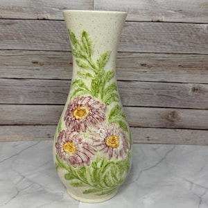 May include: A cream-colored ceramic vase with a speckled finish, featuring embossed, hand-painted pink flowers with yellow centers and green foliage. The vase has a tapered neck and a rounded body, showcasing a textured, raised floral design.