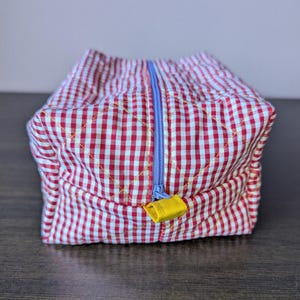 May include: A rectangular quilted pouch featuring a red and white gingham pattern with a diamond-stitched texture. The pouch includes a light blue center zipper closure and a small, bright yellow fabric tab attached to the zipper pull.