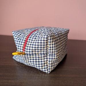 Puede incluir: Un estuche de tela acolchada con un patrón de cuadros vichy en azul marino y blanco. El bolso de forma cuadrada incluye una cremallera roja brillante en la parte superior y una pequeña lengüeta de tela amarilla unida al tirador para facilitar el acceso.