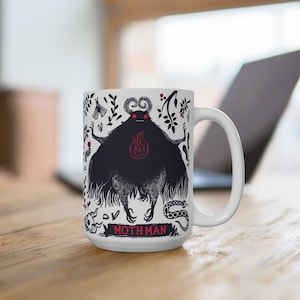 May include: White ceramic mug with a black and white illustration of a mothman with red eyes and a flame in its chest. The mug has the text "MOTH MAN" printed on the bottom.
