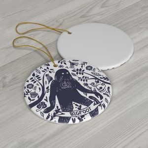 May include: White ceramic Bigfoot ornament with a navy blue illustration. The design features a Bigfoot figure with a heart, surrounded by floral and botanical elements. A gold string is attached for hanging.
