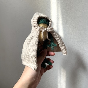 Pode incluir: Uma capa de malha bege com capuz com um brinquedo de pelúcia de sapo verde e amarelo dentro. O sapo tem um corpo verde, uma cabeça amarela e olhos pretos.