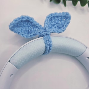 May include: A light blue, crocheted bunny ear headband accessory. The ears are attached to a band designed to fit over headphones. The accessory is made of soft yarn and is a light blue color. The background is white with green leaves.