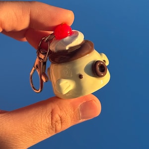 May include: A cream-colored keychain charm shaped like a small creature with a brown donut-like feature on its side. The top is decorated with a dollop of white whipped cream and a bright red cherry, attached to a silver-toned metal lobster clasp.
