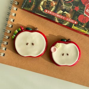 Puede incluir: Dos piezas decorativas con forma de manzana, bordes rojos y centro color crema. Una pieza presenta una pequeña oruga verde en el borde superior, mientras que la otra tiene un pequeño adorno floral rosa y amarillo. Ambas tienen dos semillas marrones.