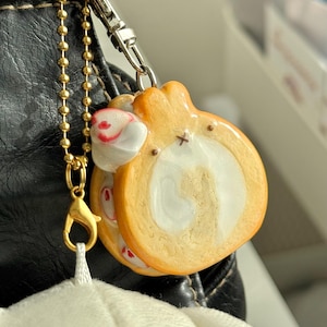May include: A miniature charm shaped like a Swiss roll cake with a golden-brown exterior and a white cream centre. The charm features a small white rabbit head with red accents attached to the side and hangs from a gold-toned metal ball chain.
