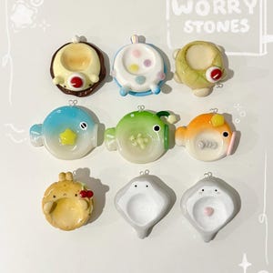 May include: Nine colorful worry stones with various designs. The top row features round stones with animal-like faces. The middle row showcases fish-shaped stones. The bottom row includes a bunny and two stingray-shaped stones. The text "Worry Stones" is visible.